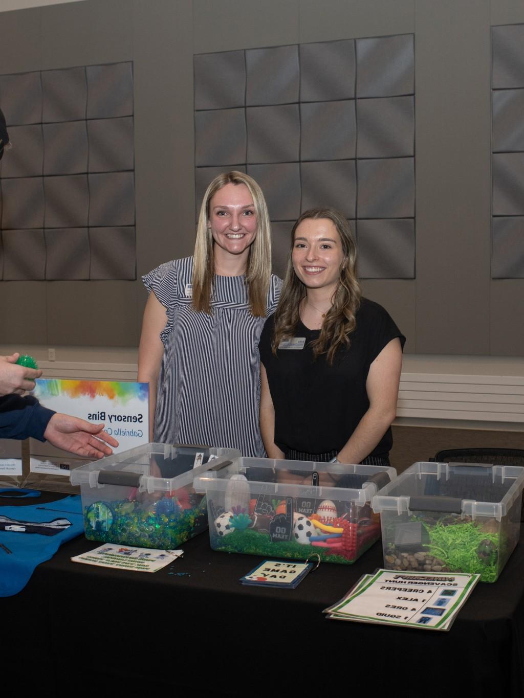 Two female students pose in front of their project, sensory bins